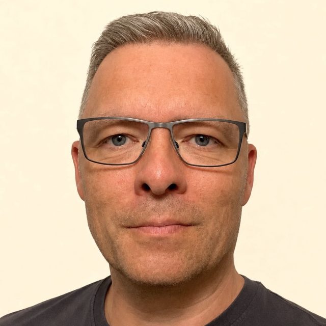 Portrait of a middle-aged man with gray hair and black rectangular glasses, wearing a dark t-shirt against a light background.